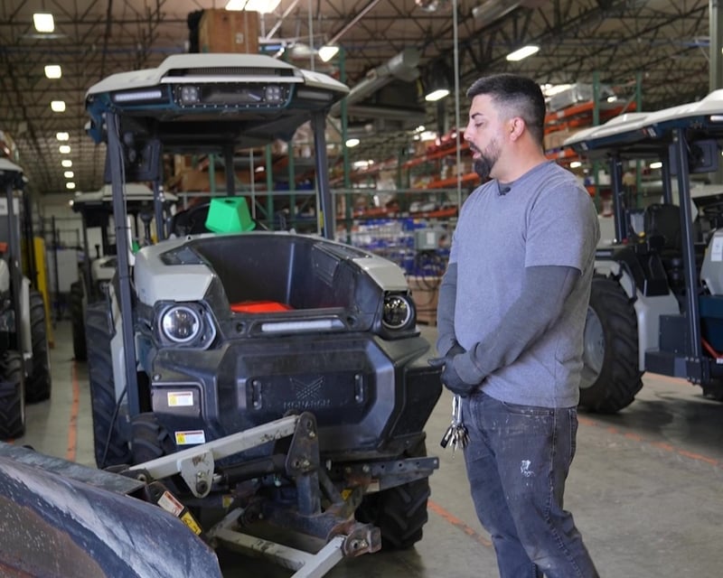Monarch Tractor Technician wearing blue t-shirt over gray shirt with long sleeves and jeans looks at the front of a MK-V electric tractor. He has short black hair and beard.