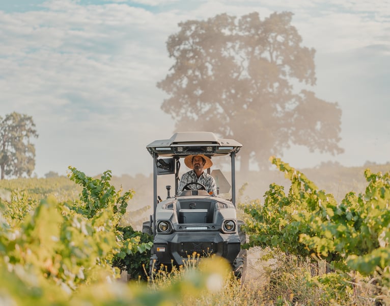 MK-V electric tractor be driven down a lush vine row, driven by a farmer in plaid shirt and straw hat.