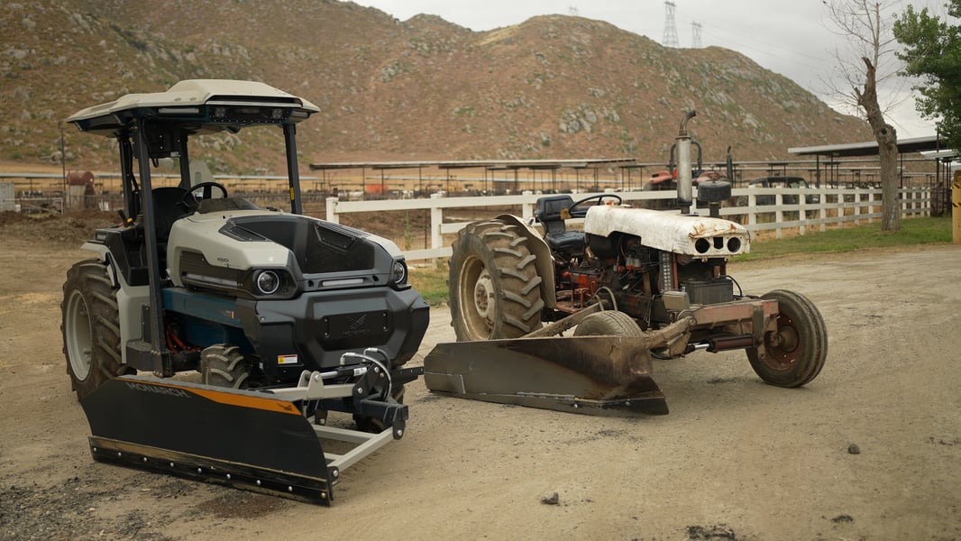 An electric tractor and a diesel-powered tractor with dairy equipment — a feed push blade implement — sit side-by-side.