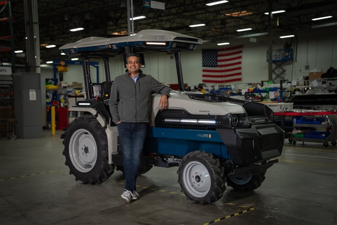 Monarch CEO, Praveen Penmetsa, stands next to the gray and teal Monarch Tractor. His elbow is casually resting on the tractor. A U.S. flag hangs on the wall in the background.