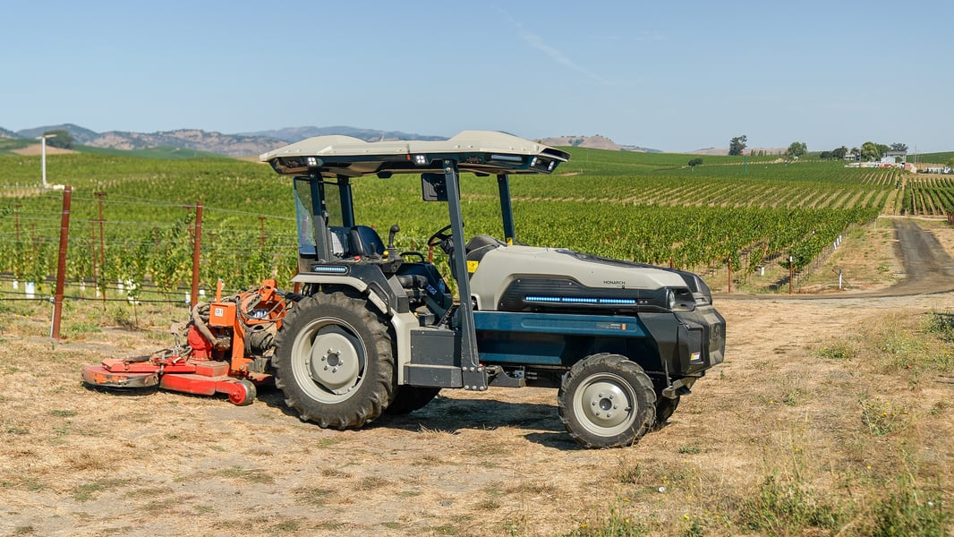 MK-V electric tractor pulling an implement in a vineyard. Background is rolling vineyard countryside with California's golden hills in the far distance against a pale blue sky. 